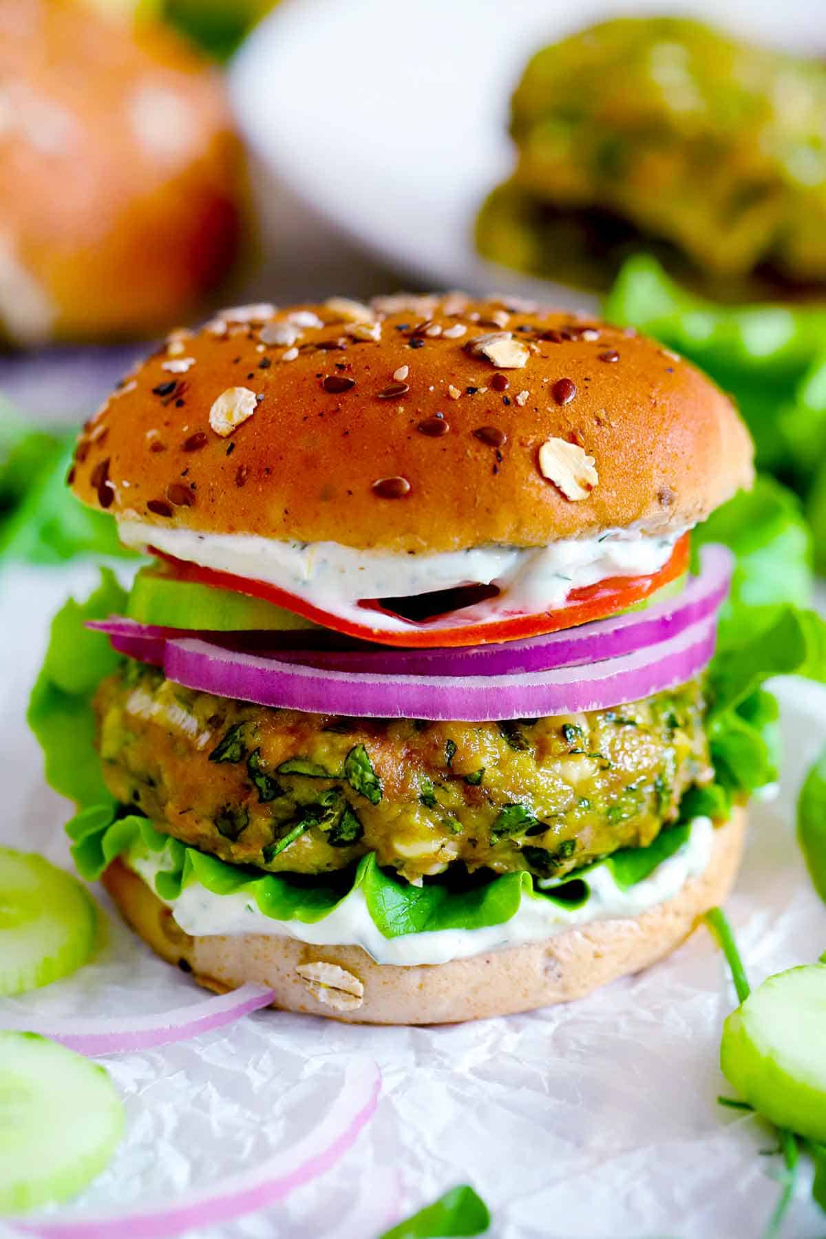 Close up side-view of a spanakopita turkey burger with red onions, tomatoes, cucumbers, lettuce, and yogurt sauce on a whole grain bun.