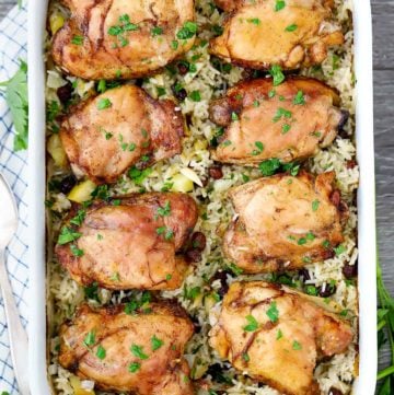 A casserole dish with chicken thighs baked on top of rice, apples, and raisins on a plaid towel.