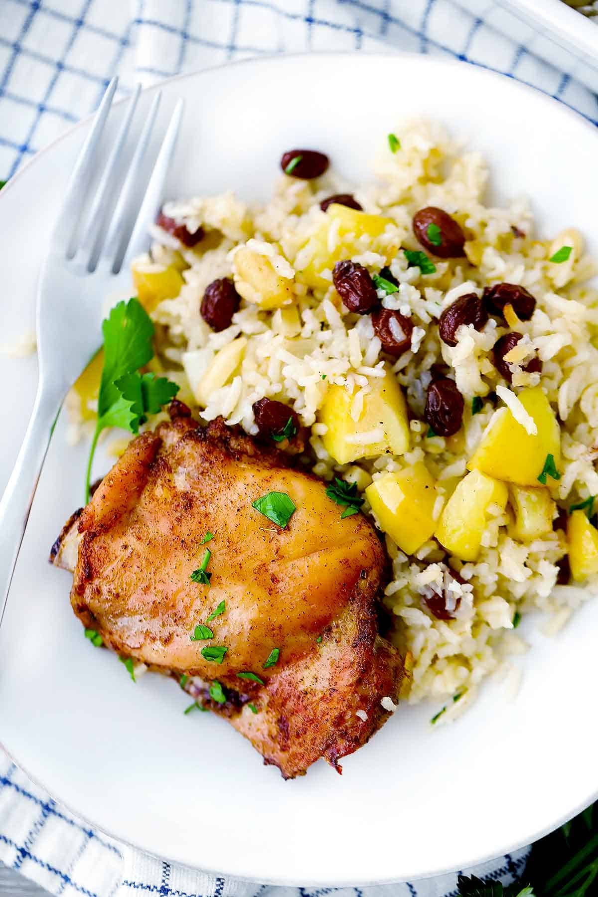 A white plate with a chicken thigh and rice with raisins and apples.