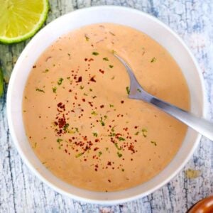 Square photo of overhead view of a bowl of homemade spicy mayo with a spoon in it, topped with lime zest and red pepper.