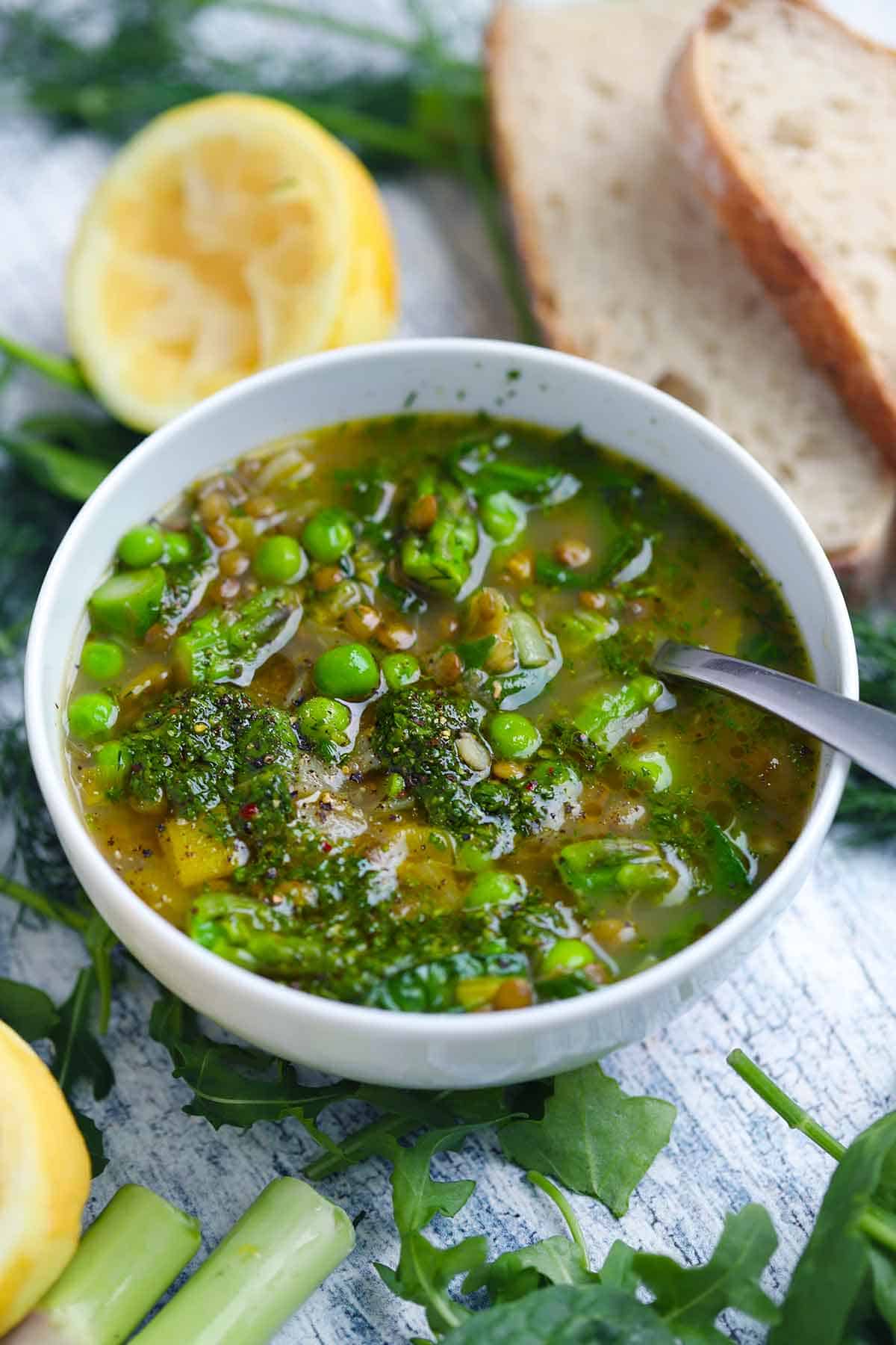 Angled view of a bowl of spring lentil vegetable soup with lemons and bread scattered around it.