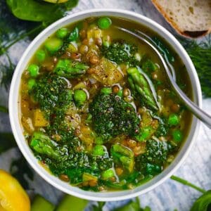 Square photo of an overhead view of spring lentil vegetable soup with bright green veggies and herb pistou spooned on top.