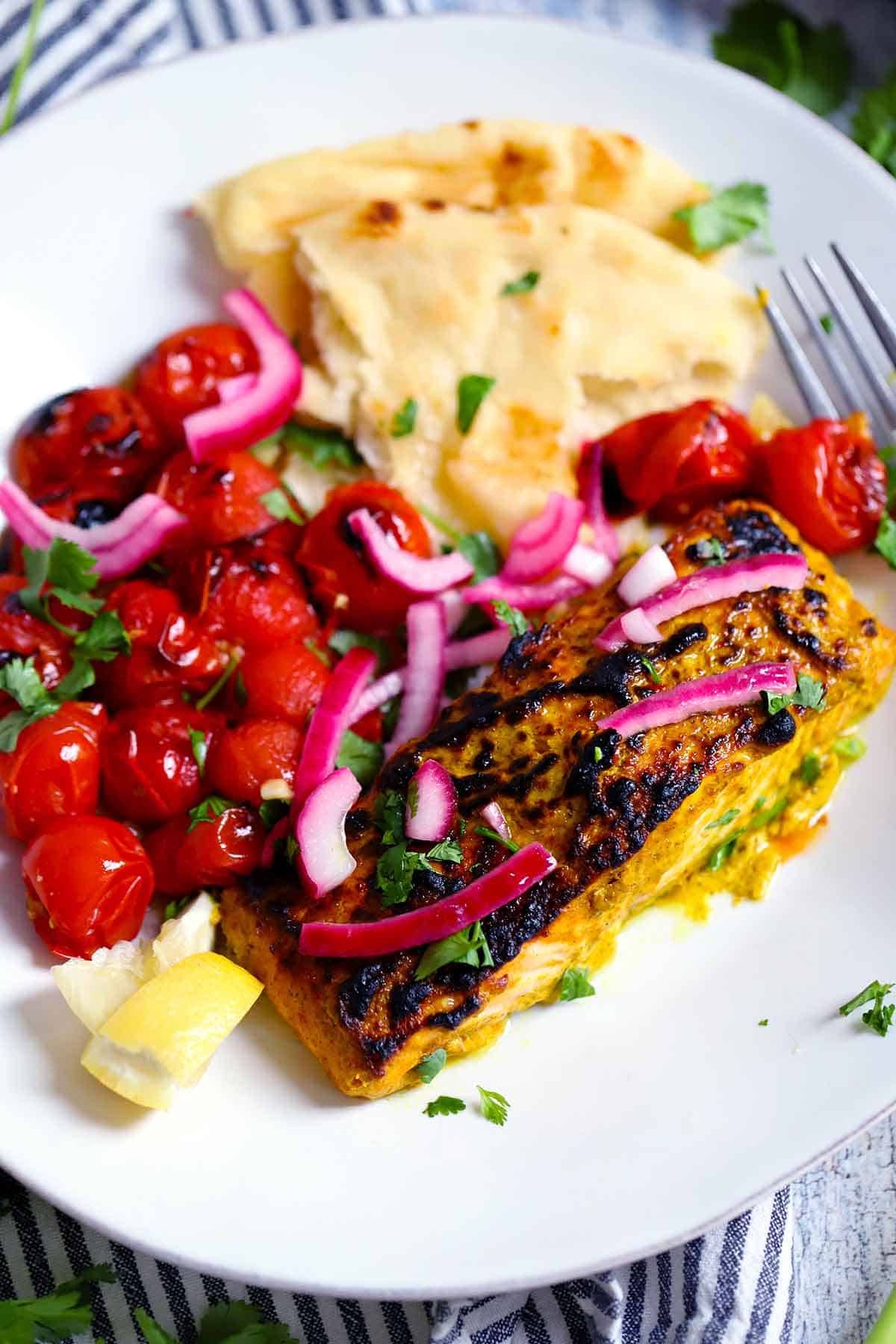 Tandoori salmon and cherry tomatoes on a plate topped with cilantro and pickled red onions with naan on the side.