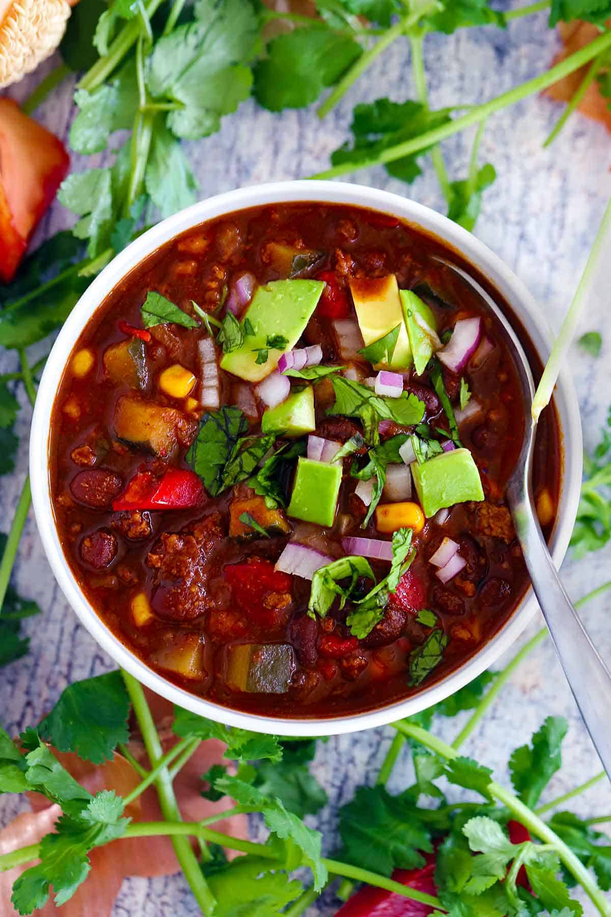 Overhead photo of a bowl of turkey chili with veggies and black beans topped with avocado, cilantro, and red onion, with ingredient scraps scattered around.