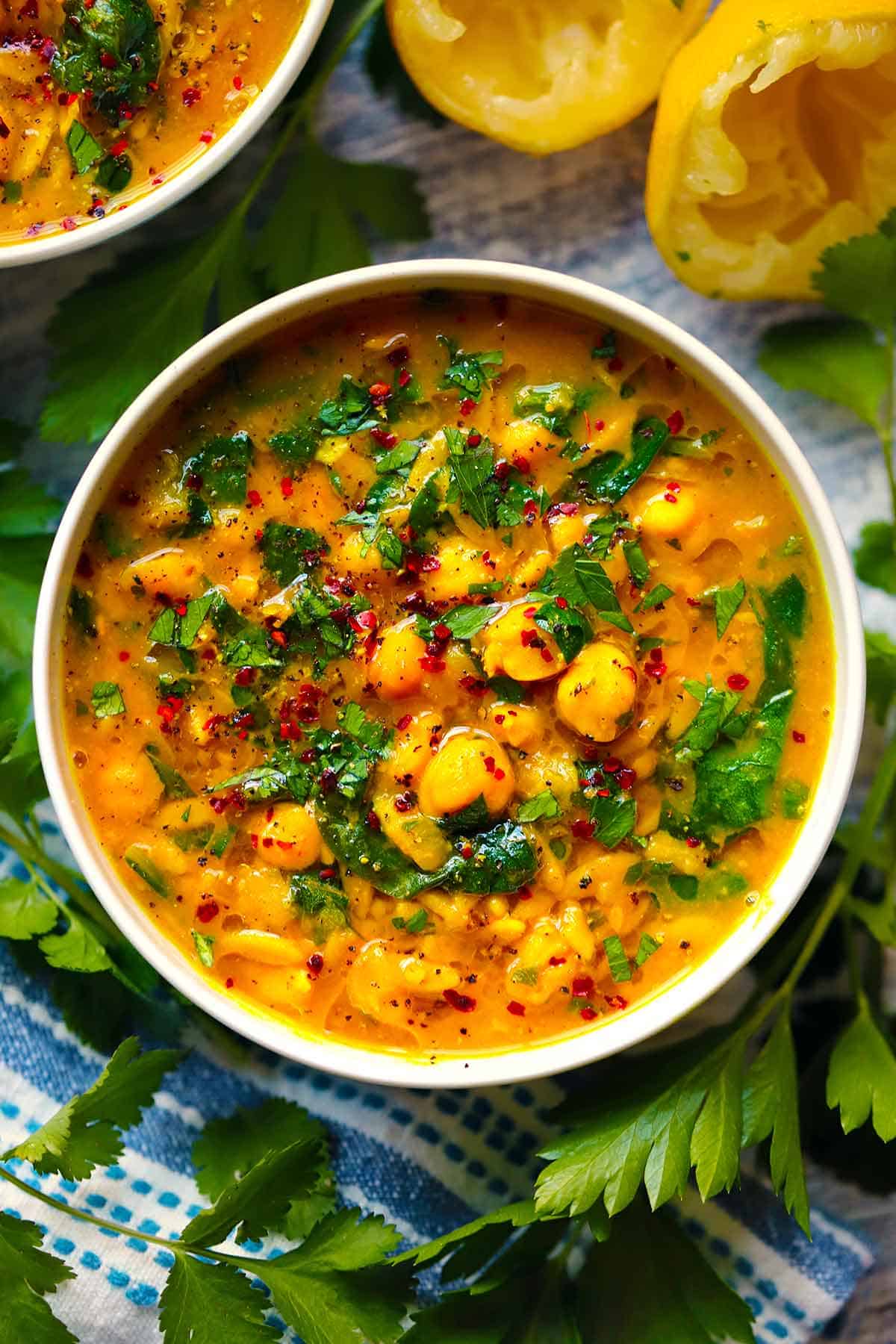 Overhead view of a bowl of turmeric soup with orzo, chickpeas, spinach, and lemon, with aleppo pepper and olive oil on top, with lemon and parsley scraps scattered around.