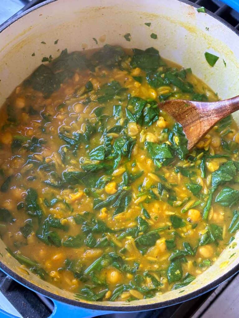 Finished turmeric soup with chickpeas, orzo, spinach, and lemon in a Dutch oven pot with a wooden spoon.