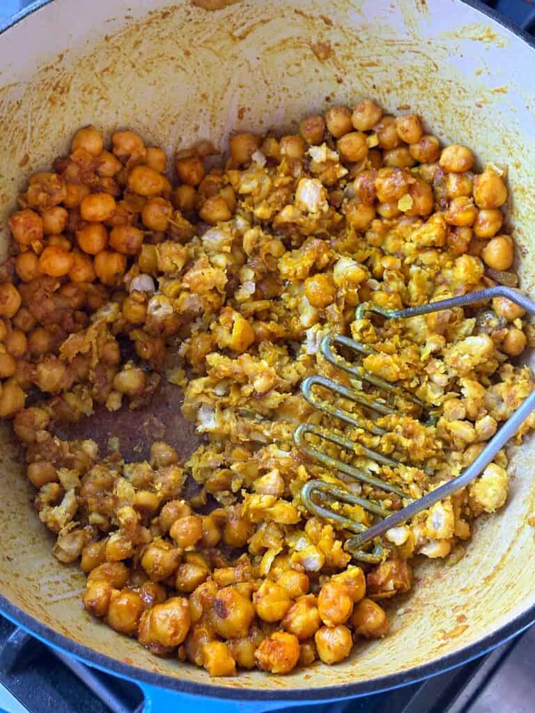 Using a potato masher to mash chickpeas after sautéing in spices in a pot.
