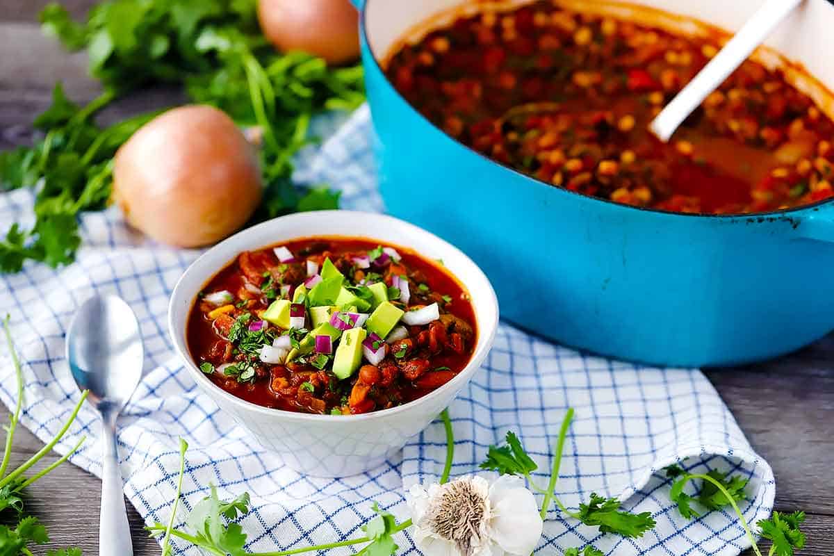 A bowl of vegetarian chili in front of a blue dutch oven on a white checkered towel.