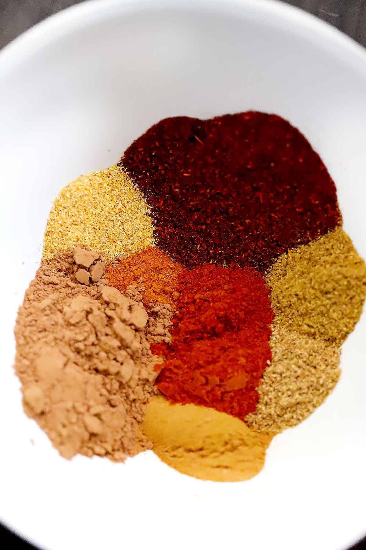 chili powder, garlic powder, cocoa powder, cayenne pepper, garlic powder, cumin, coriander, cinnamon, and smoked paprika in a white bowl.